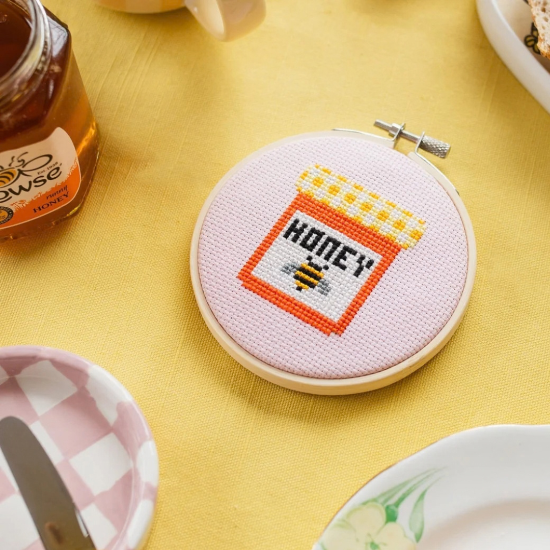 Completed honey jar cross stitch displayed in a bamboo hoop on a yellow table with colourful tableware.