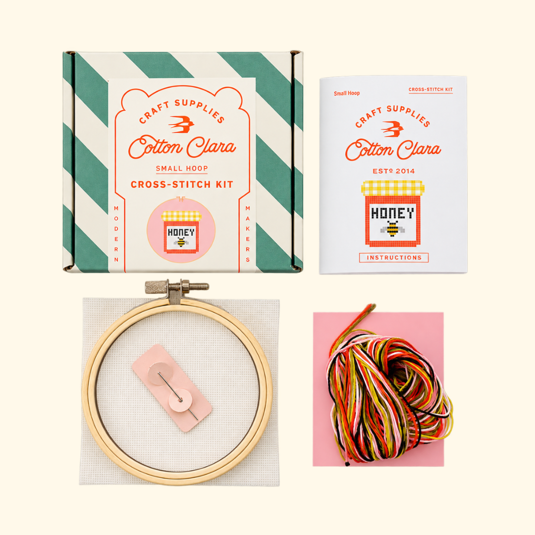 Contents of the Cotton Clara Honey Jar Cross Stitch Kit including bamboo hoop, pink Aida fabric, embroidery threads, needle and instructions.