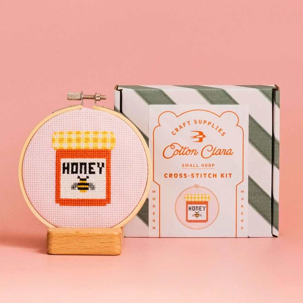 Finished honey jar cross stitch in a bamboo hoop displayed beside the Cotton Clara kit box on a pink background.