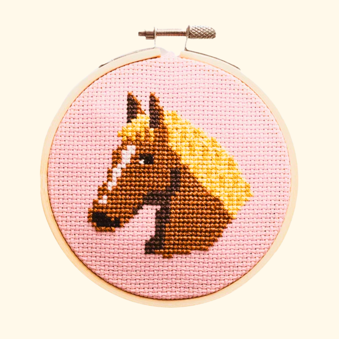 Finished horse cross stitch design in brown and golden yellow thread, displayed in a bamboo embroidery hoop on pink Aida fabric.