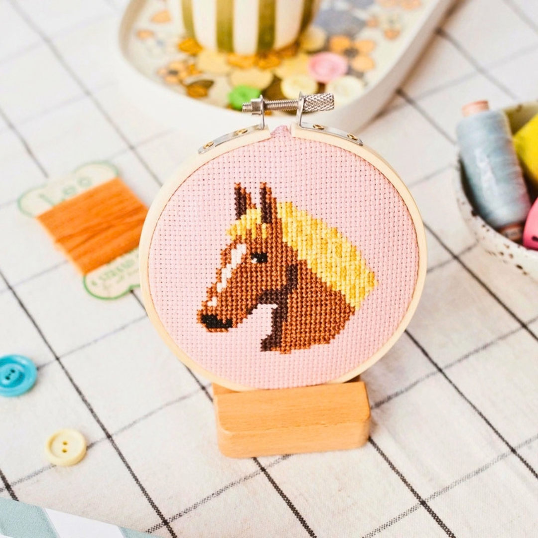 Completed horse cross stitch displayed in a bamboo hoop with colourful craft supplies in the background.
