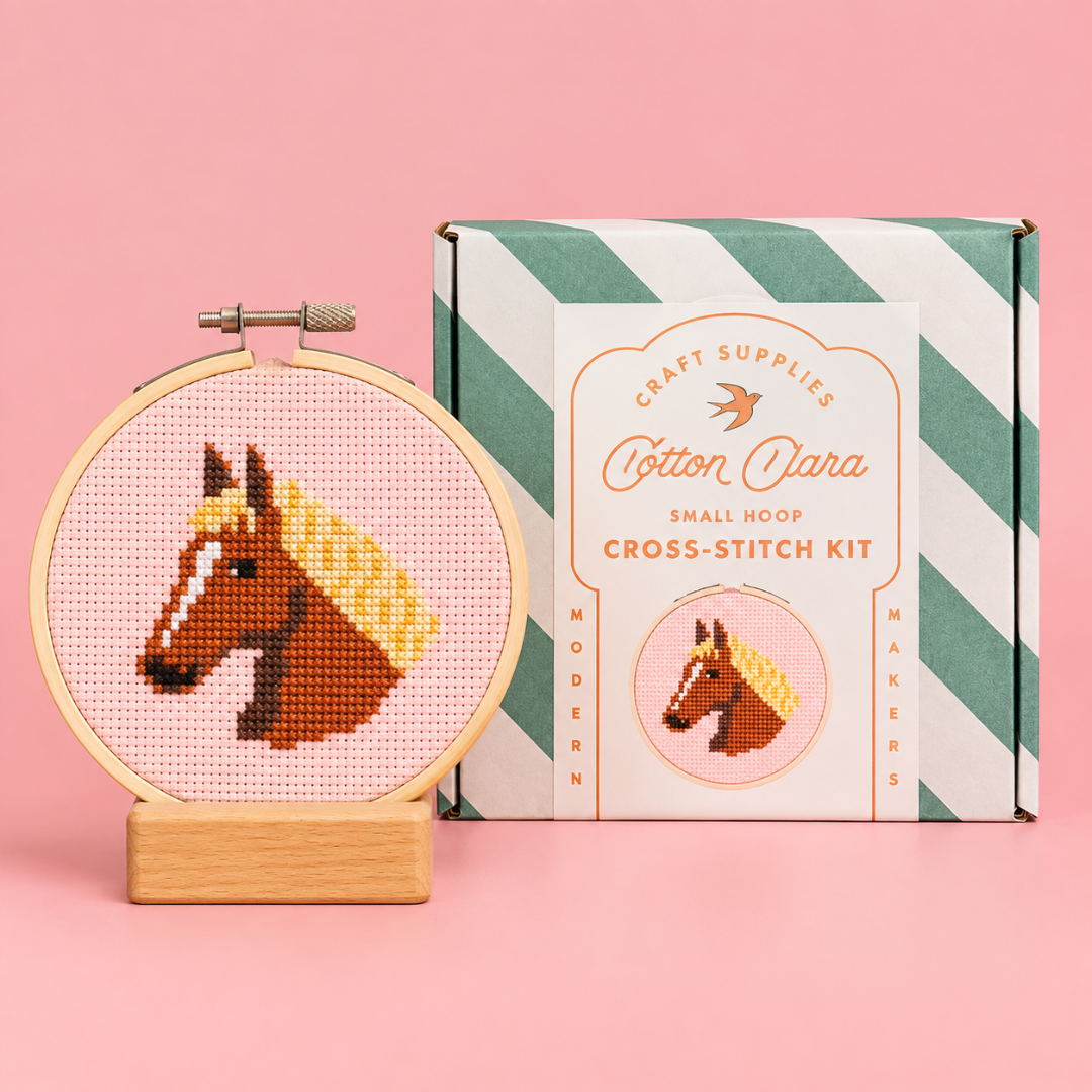 Finished horse cross stitch in a bamboo hoop displayed beside the Cotton Clara kit box on a pink background.
