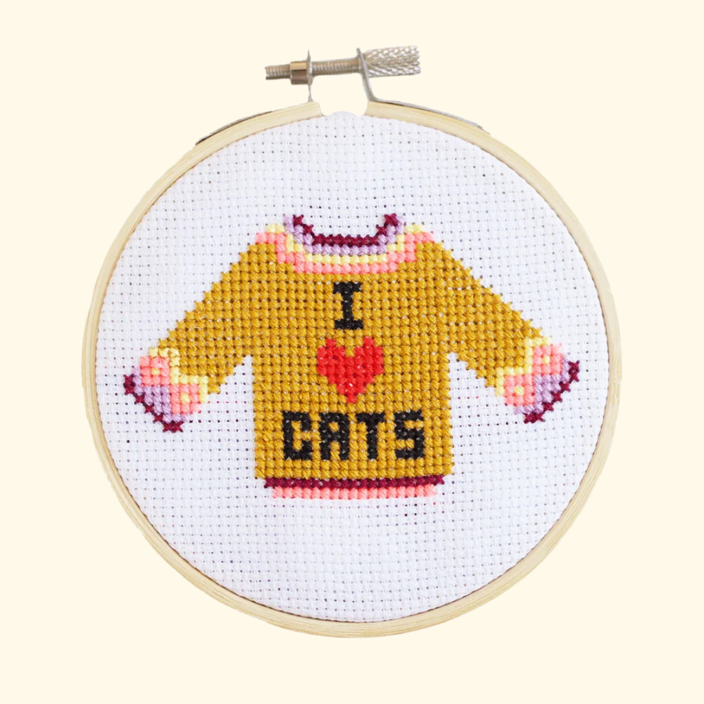 Finished I Love Cats cross stitch featuring a mustard yellow jumper with pink details and “I ♥ CATS” lettering, displayed in a bamboo embroidery hoop.