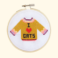 Finished I Love Cats cross stitch featuring a mustard yellow jumper with pink details and “I ♥ CATS” lettering, displayed in a bamboo embroidery hoop.
