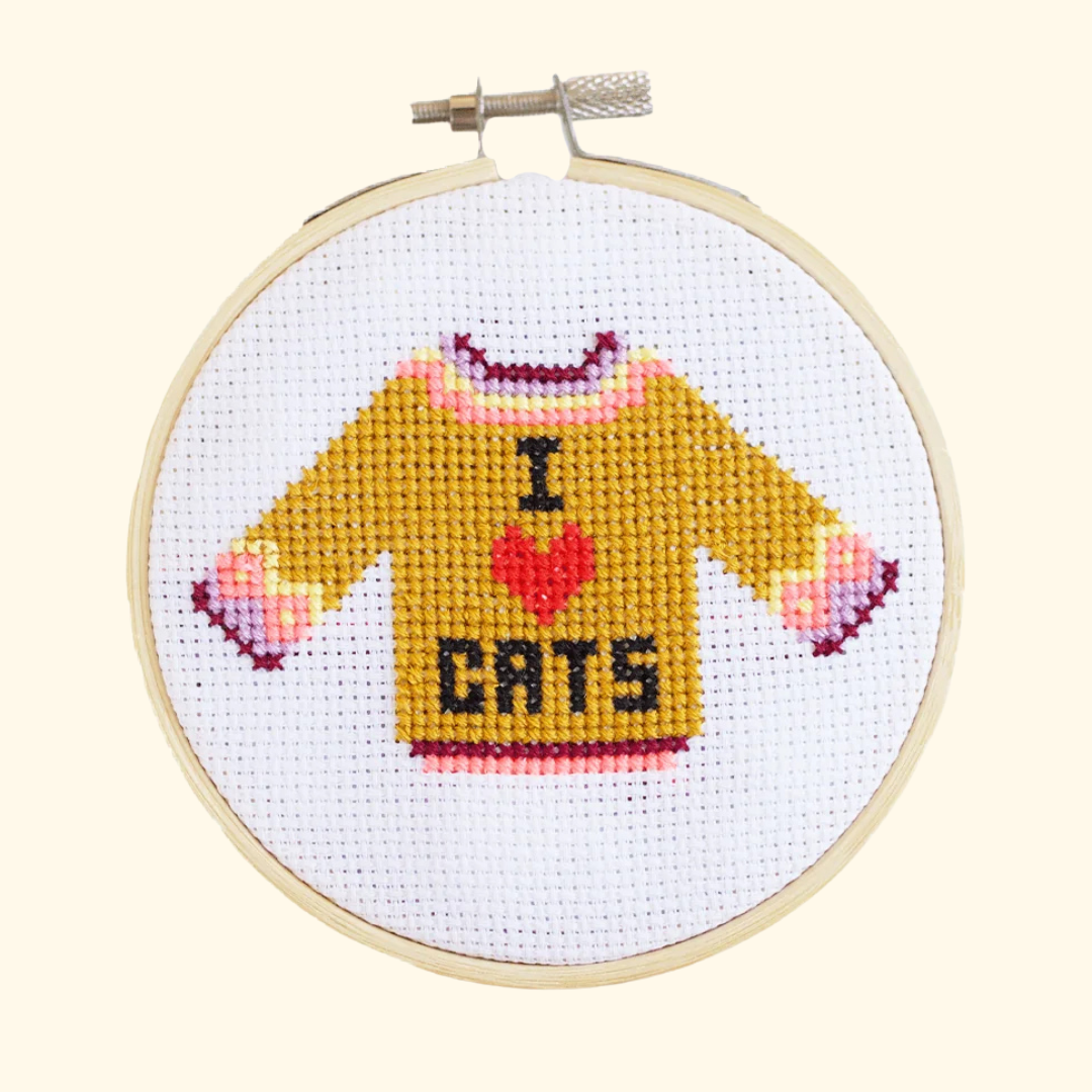 Finished I Love Cats cross stitch featuring a mustard yellow jumper with pink details and “I ♥ CATS” lettering, displayed in a bamboo embroidery hoop.