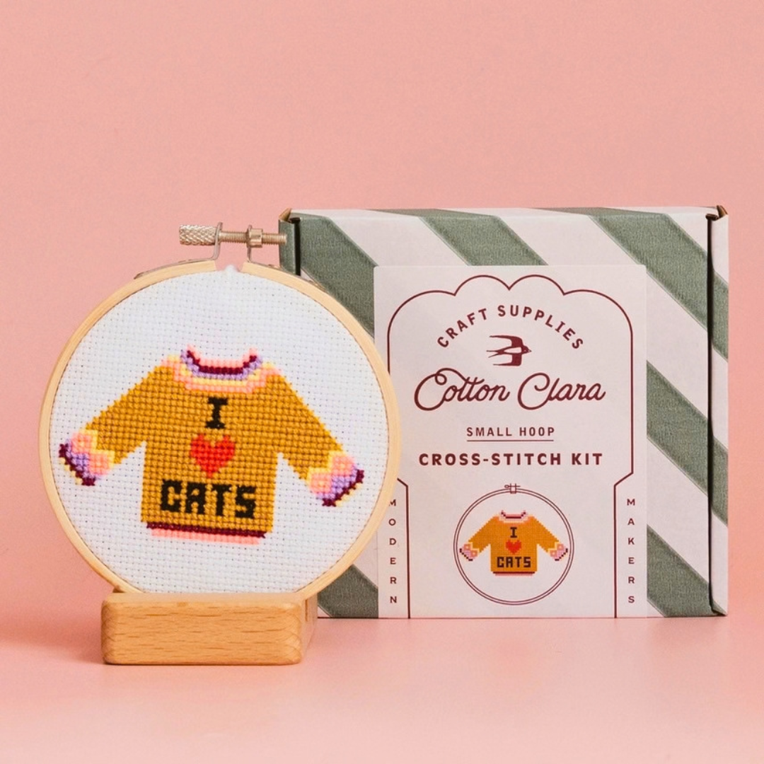 Completed I Love Cats jumper cross stitch displayed in a bamboo hoop beside the Cotton Clara kit box on a pink background.