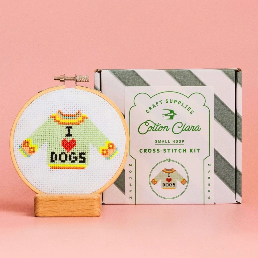 Finished I Love Dogs jumper cross stitch displayed in a bamboo hoop beside the Cotton Clara kit box on a pink background.