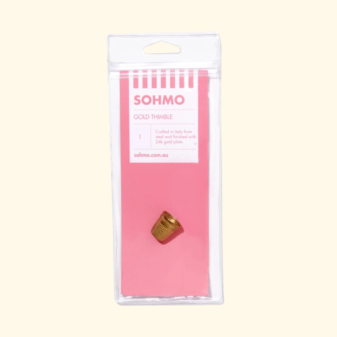 A gold metal thimble inside a clear plastic hanging package with a pink backing card labeled SOHMO GOLD THIMBLE, shown on a light cream background.