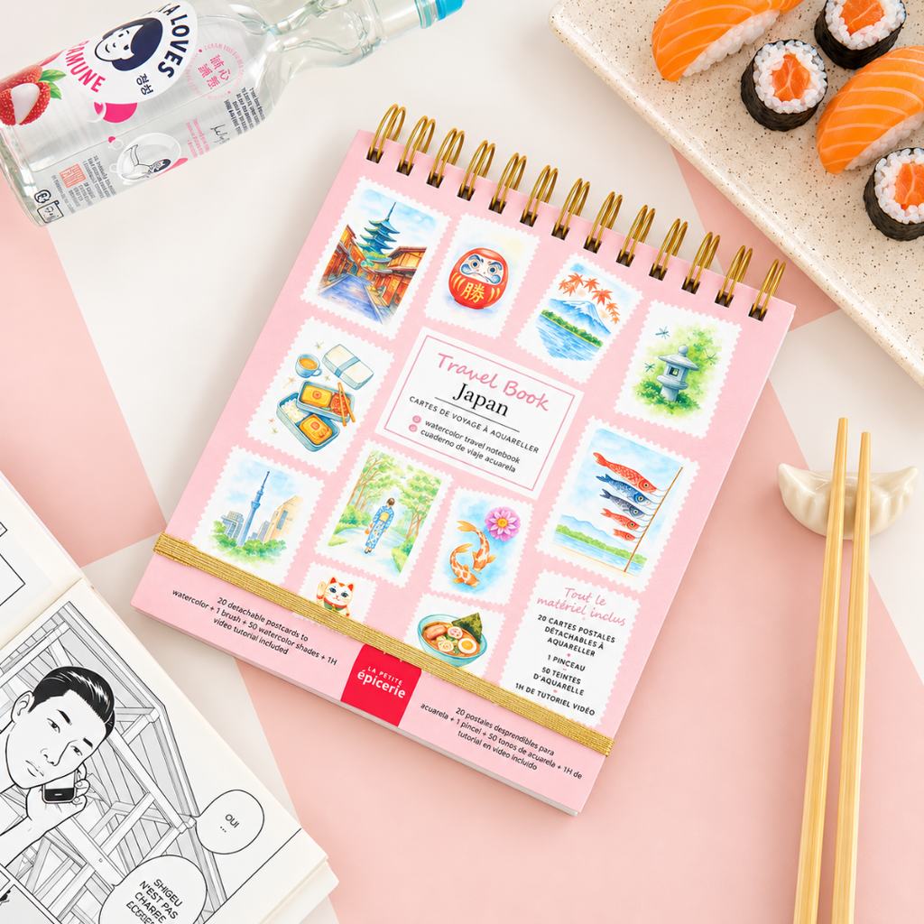 Japan Watercolour Travel Book by La Petite Epicerie displayed on a pink surface alongside Japanese-inspired artwork and painting accessories, featuring colourful illustrations on the book cover.