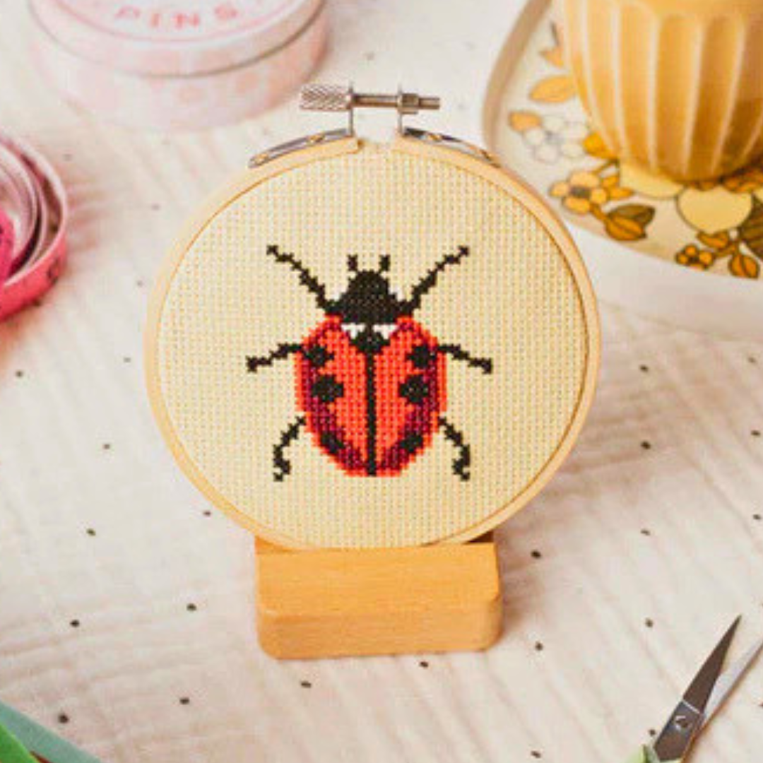 Finished ladybird cross stitch displayed in a bamboo embroidery hoop on a wooden stand.