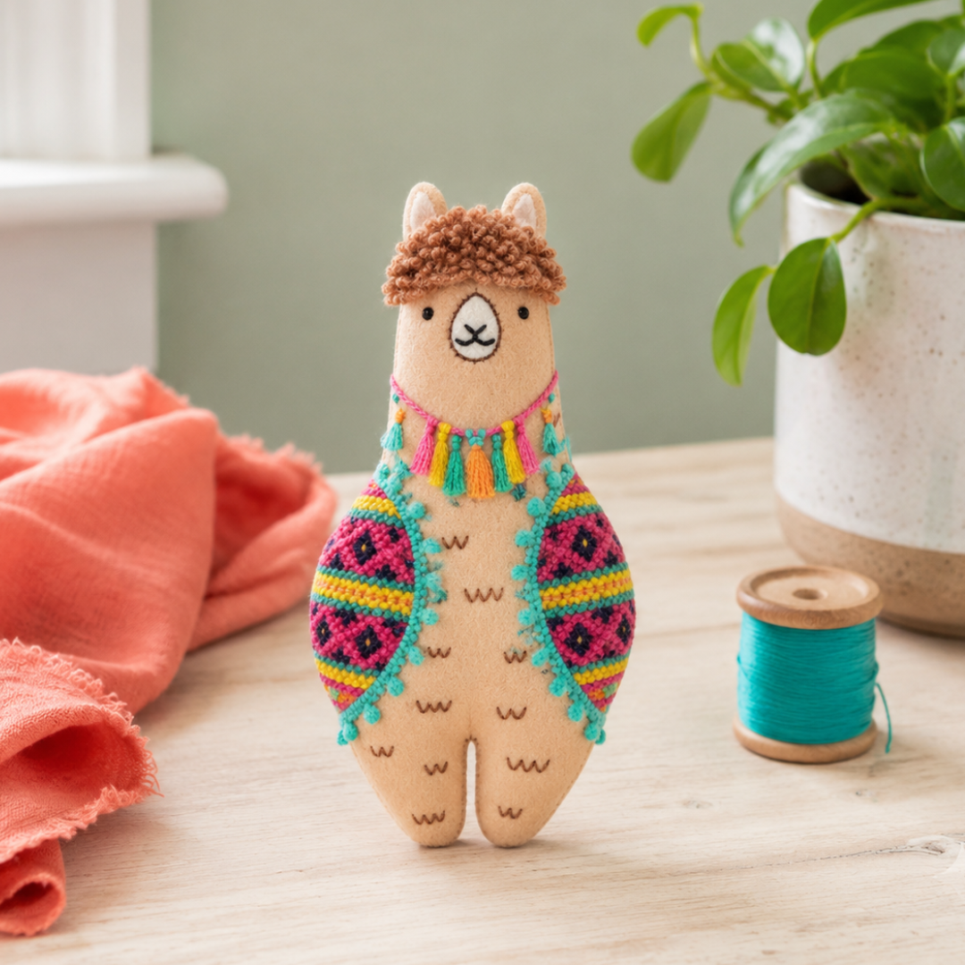 Finished Kiriki Press llama embroidery doll displayed with colourful fabric, thread and a potted plant.