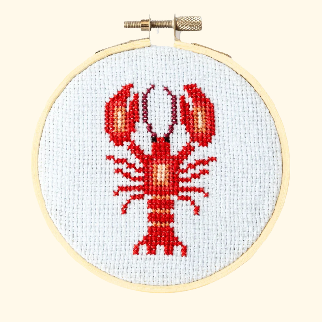 Finished red lobster cross stitch displayed in a wooden embroidery hoop on white Aida fabric.