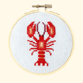 Finished red lobster cross stitch displayed in a wooden embroidery hoop on white Aida fabric.