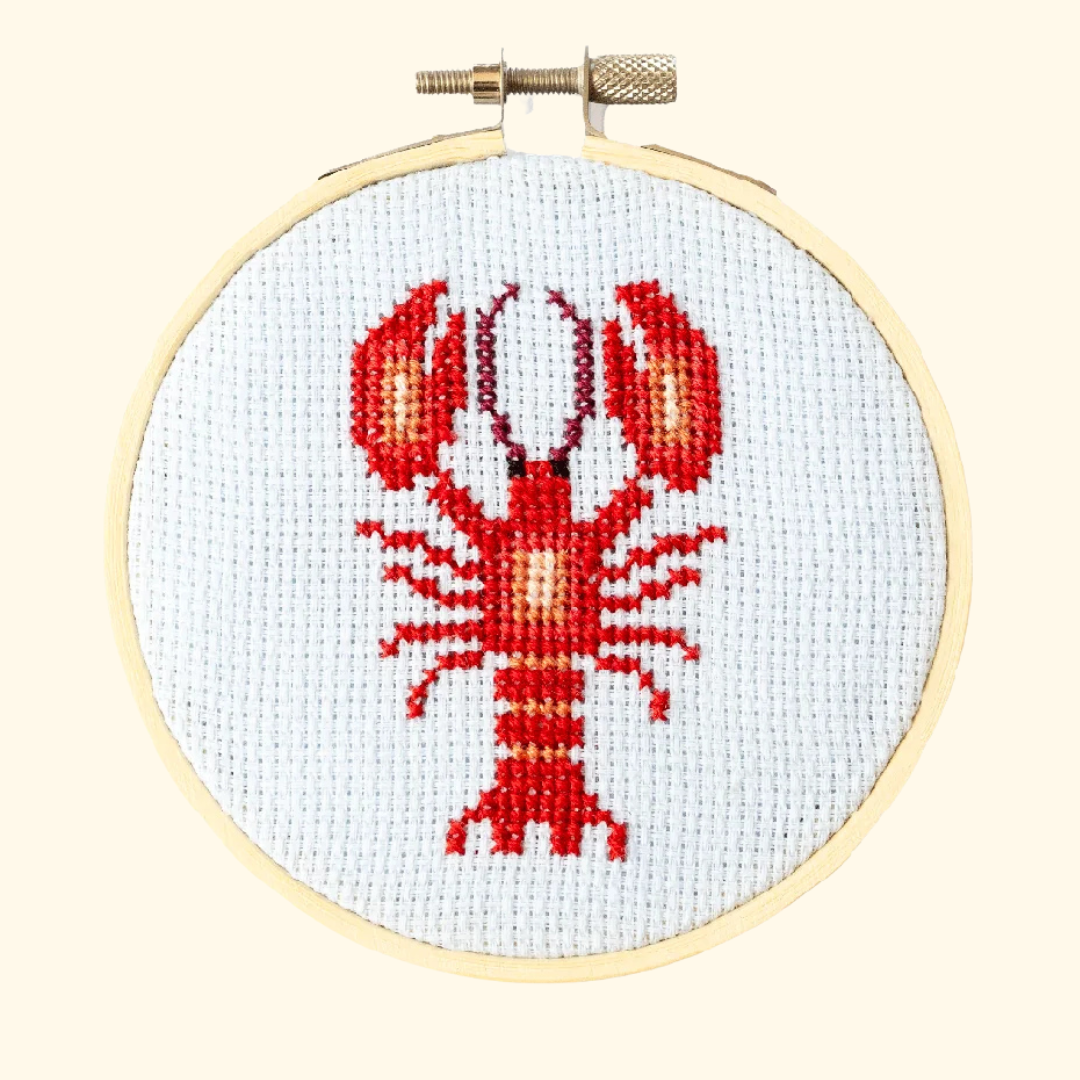 Finished red lobster cross stitch displayed in a wooden embroidery hoop on white Aida fabric.