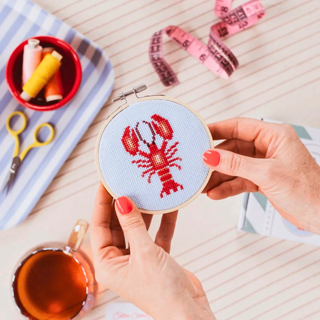 Lobster cross stitch being stitched by hand with pink fabric, embroidery threads and craft supplies surrounding the hoop.