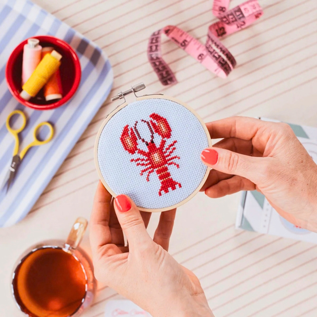 Lobster cross stitch being stitched by hand with pink fabric, embroidery threads and craft supplies surrounding the hoop.