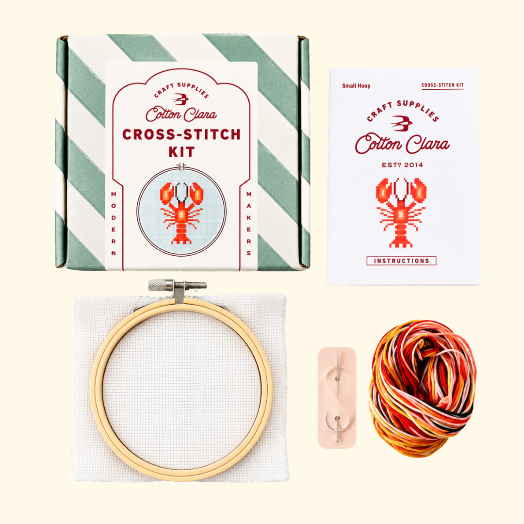 Contents of the Cotton Clara Lobster Cross Stitch Kit including hoop, Aida fabric, red embroidery thread, needle and instructions.