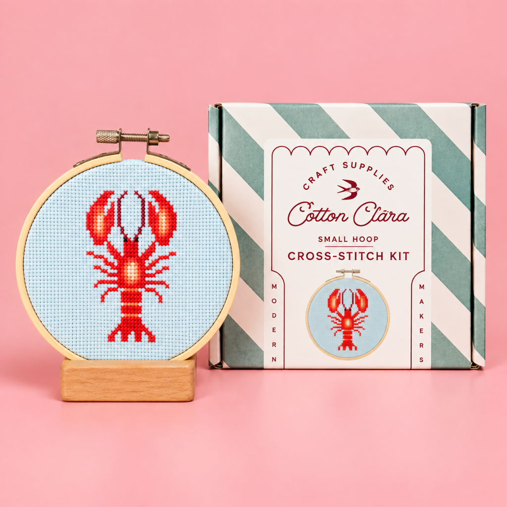 Completed red lobster cross stitch in a wooden hoop displayed beside the Cotton Clara kit box on a pink background.