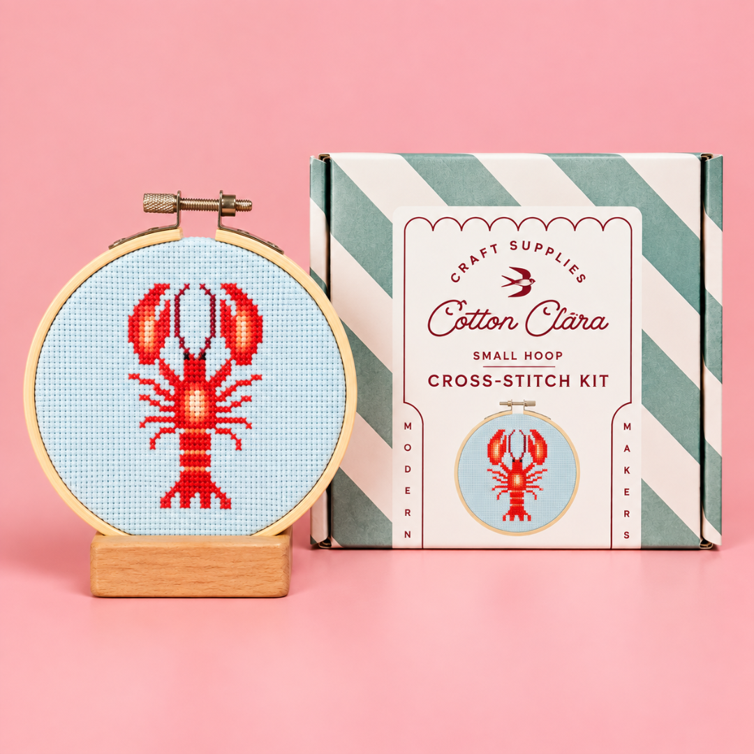 Completed red lobster cross stitch in a wooden hoop displayed beside the Cotton Clara kit box on a pink background.