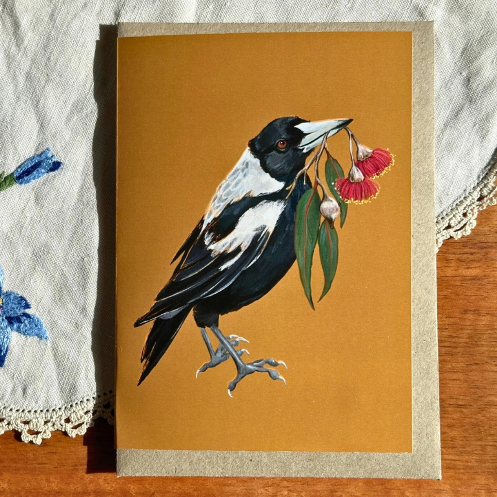 Magic Magpie greeting card styled on a wooden surface with lace and blue flowers, featuring an Australian magpie carrying a flowering gum branch.