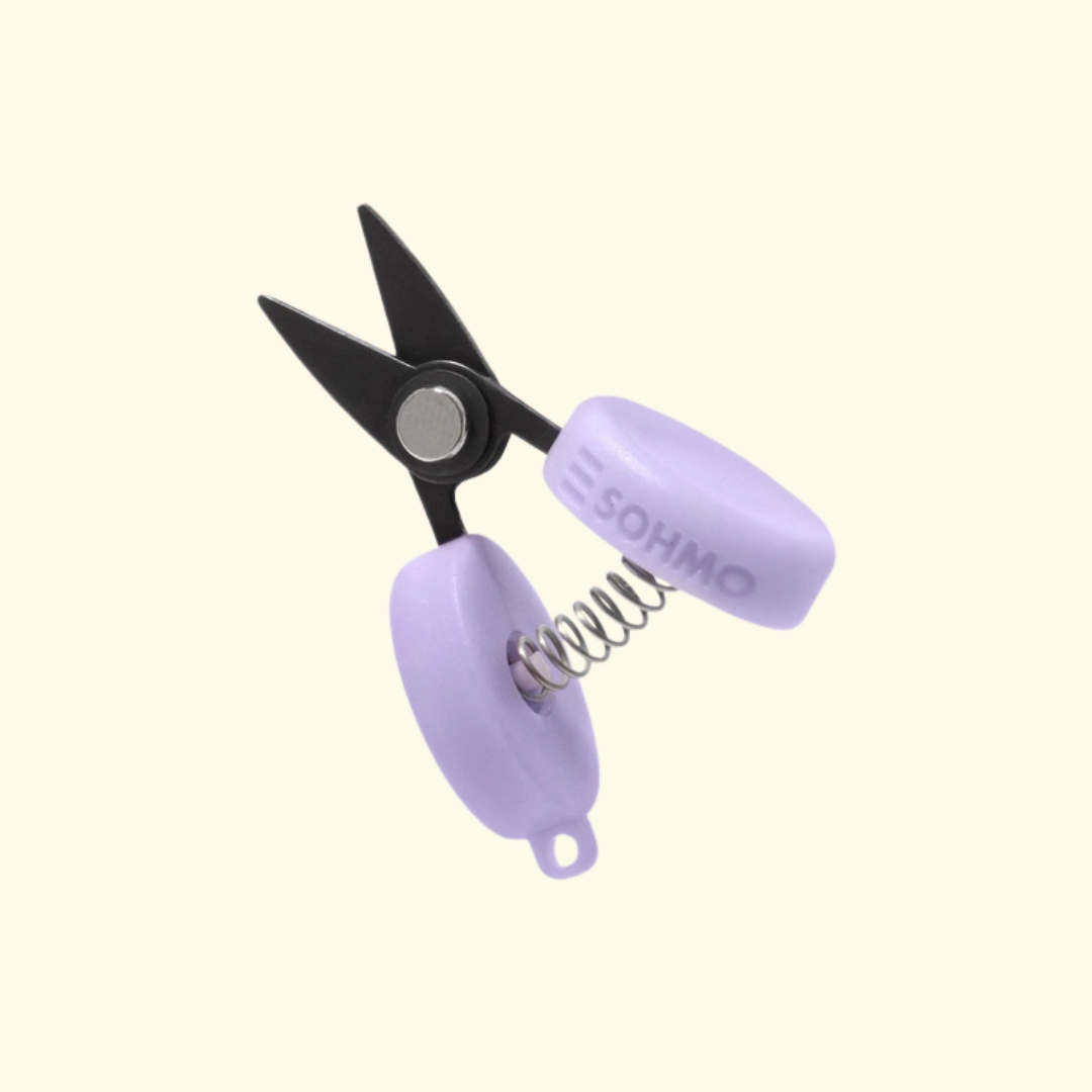 A pair of mini spring scissors with black metal blades and light purple plastic handles, featuring a visible spring and the brand name 'SOHMO' printed on one handle, set against a solid cream background.