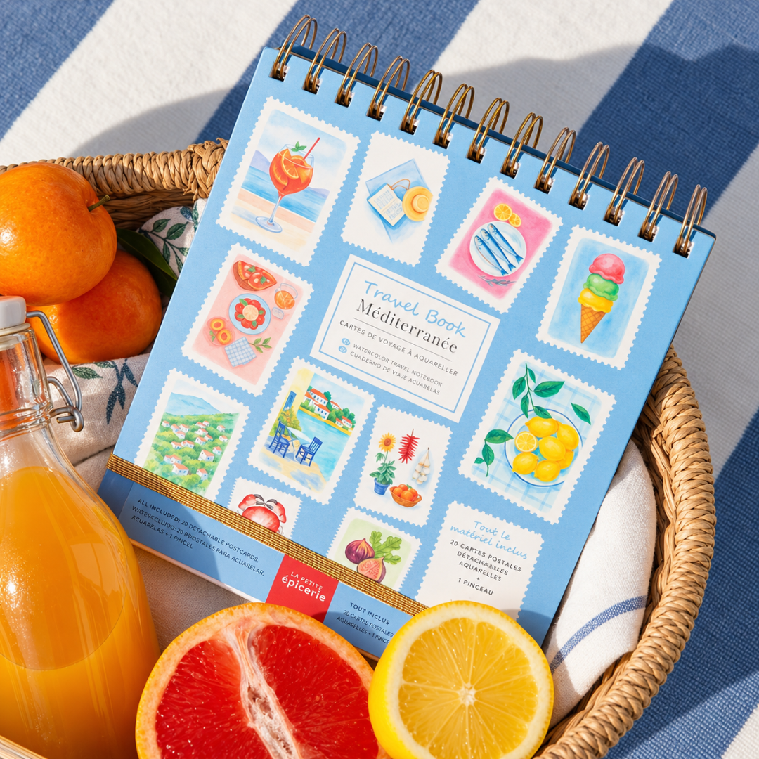 Mediterranean Watercolour Travel Book by La Petite Epicerie displayed inside a blue striped beach bag alongside fresh oranges, featuring colourful Mediterranean-inspired illustrations on the cover.