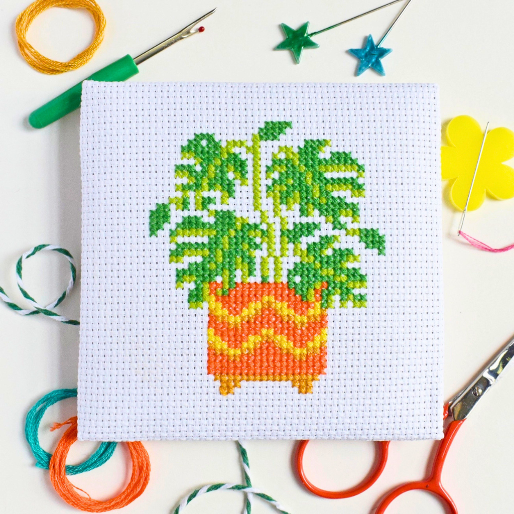 Completed monstera cross stitch design styled with colourful craft supplies on a blue and white striped surface.
