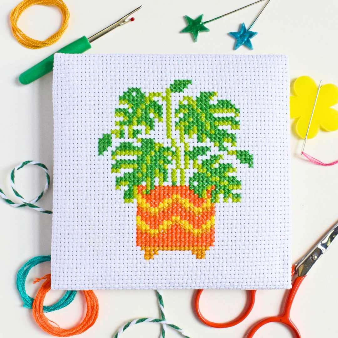 Completed monstera cross stitch design styled with colourful craft supplies on a blue and white striped surface.