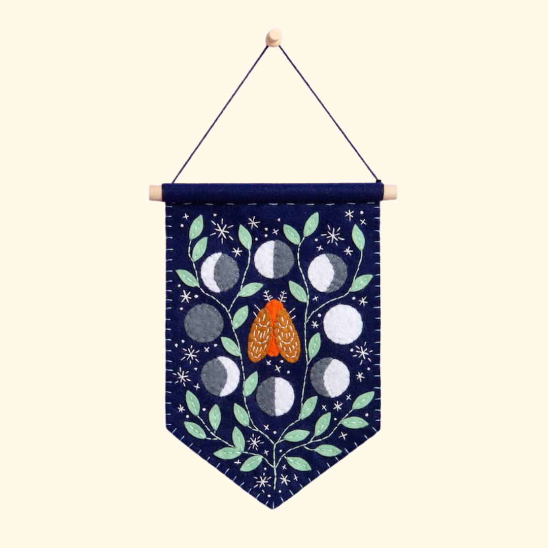 Felt wall hanging featuring an orange moth surrounded by moon phases, leaves and stars on a dark blue background