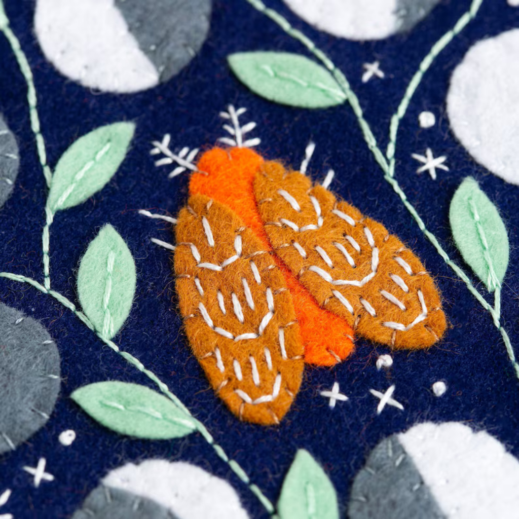 Close up of orange felt moth with embroidered details surrounded by leaves and moon phases