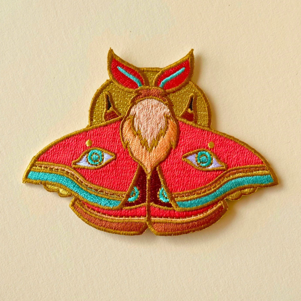 Close-up of Ashlea Bechaz Illustration moth embroidered iron on patch showcasing detailed red, green and golden yellow wings with intricate symmetrical patterns against a beige fabric background.