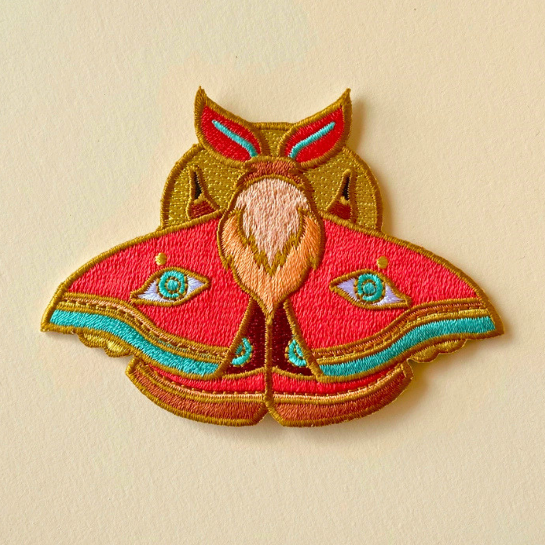Close-up of Ashlea Bechaz Illustration moth embroidered iron on patch showcasing detailed red, green and golden yellow wings with intricate symmetrical patterns against a beige fabric background.