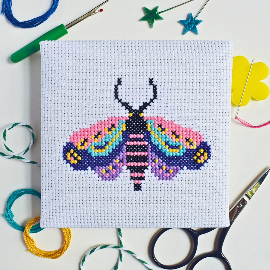 Completed moth cross stitch design styled on a green and white striped craft table.
