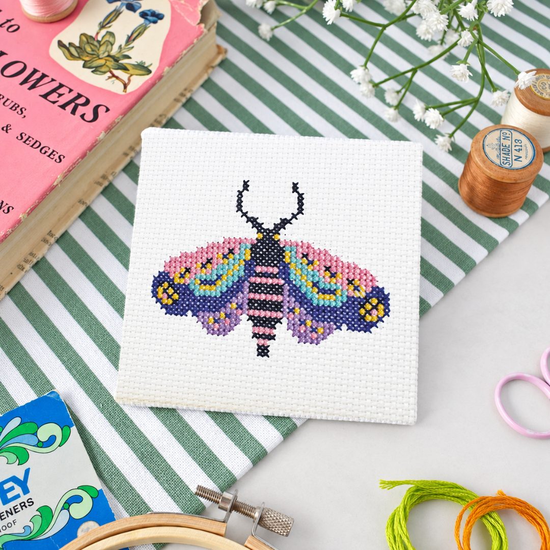 Colourful moth cross stitch design surrounded by scissors, thread, a needle, and an embroidery hoop.