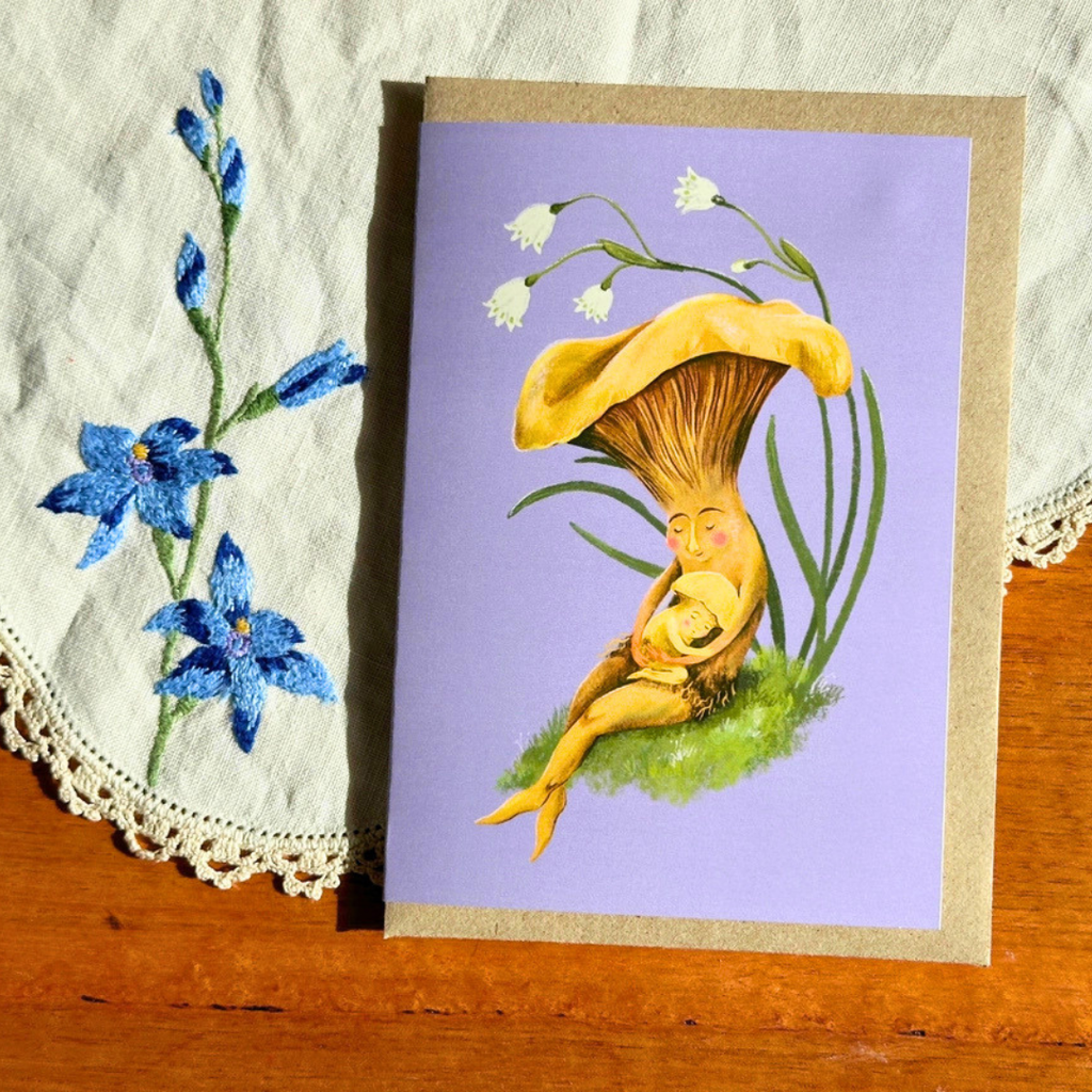 Mushroom Snuggles greeting card styled on a wooden surface with lace and blue flowers, featuring a whimsical mother and baby mushroom illustration.
