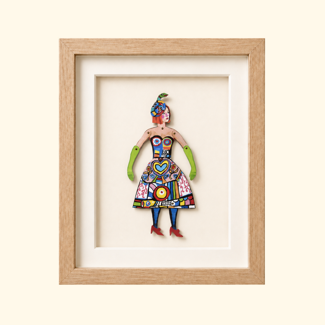 Framed Niki de Saint Phalle paper puppet wearing a brightly patterned dress and sculptural snake headdress.