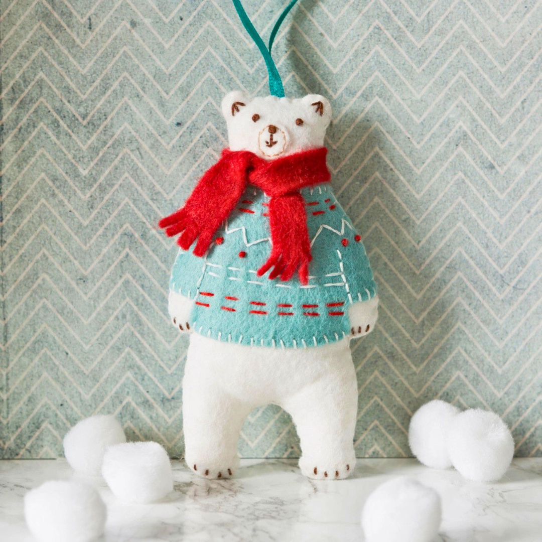 Handmade polar bear felt figure wearing a red scarf and patterned jumper, styled with white pom poms against a winter backdrop.