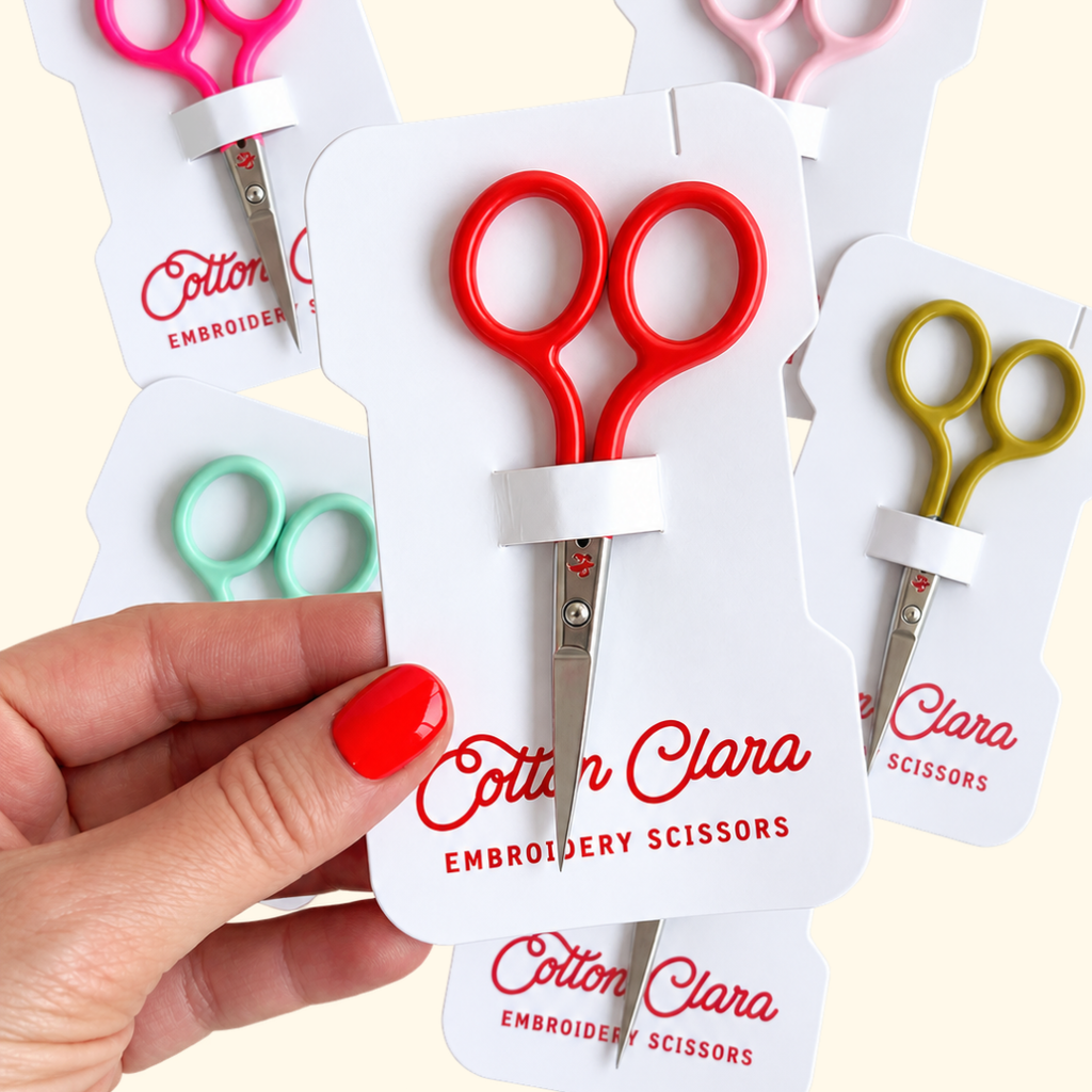 Poppy red Cotton Clara embroidery scissors held in hand, displayed alongside other colourful embroidery scissors in mint green, pink and gold.