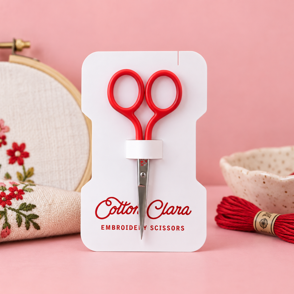 Poppy red Cotton Clara embroidery scissors on branded packaging, styled with an embroidery hoop and colourful craft supplies on a pink background.