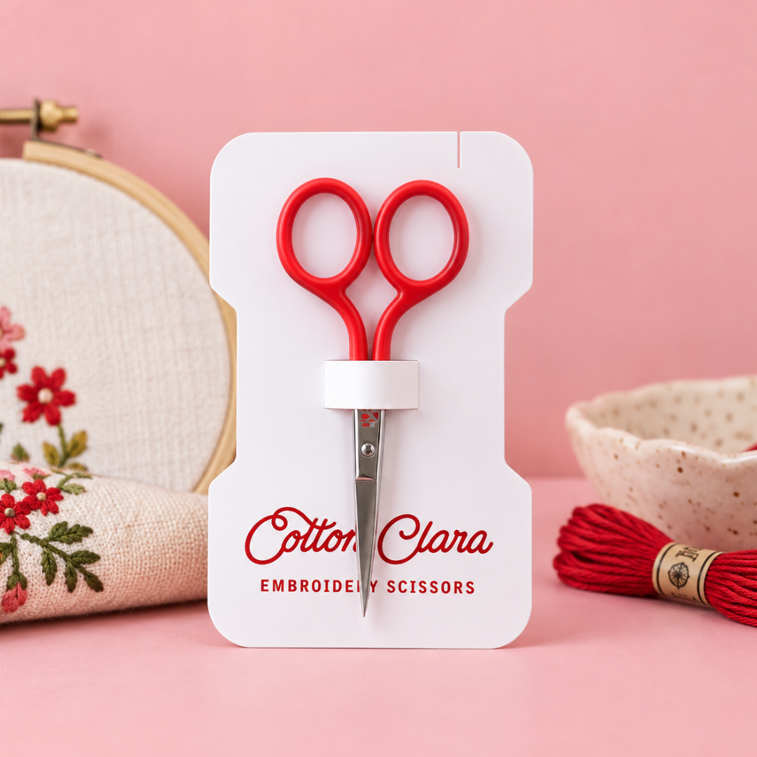 Poppy red Cotton Clara embroidery scissors on branded packaging, styled with an embroidery hoop and colourful craft supplies on a pink background.