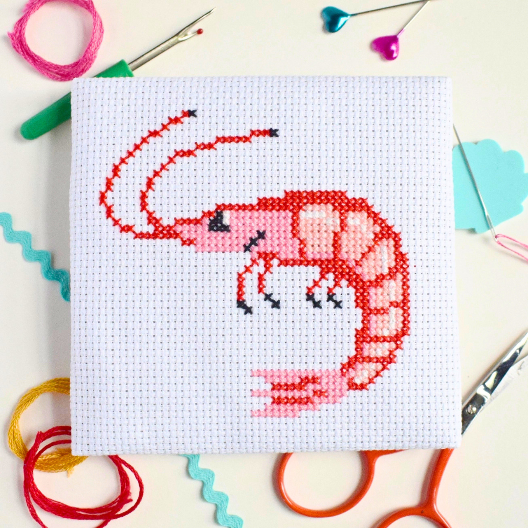 Red prawn cross stitch design surrounded by scissors, thread, a needle and colourful paper.