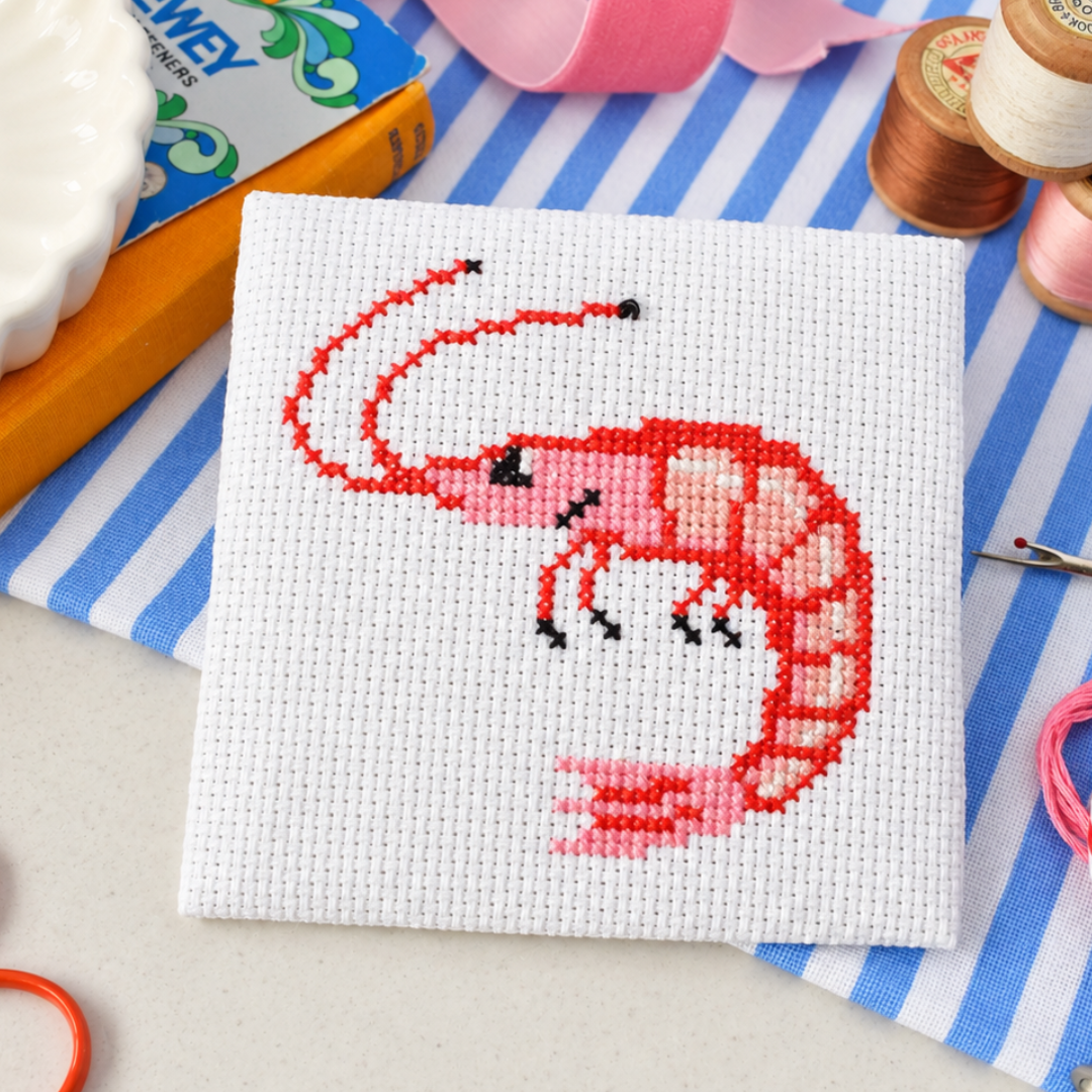 Completed prawn cross stitch design styled with colourful craft supplies on a blue and white striped surface.