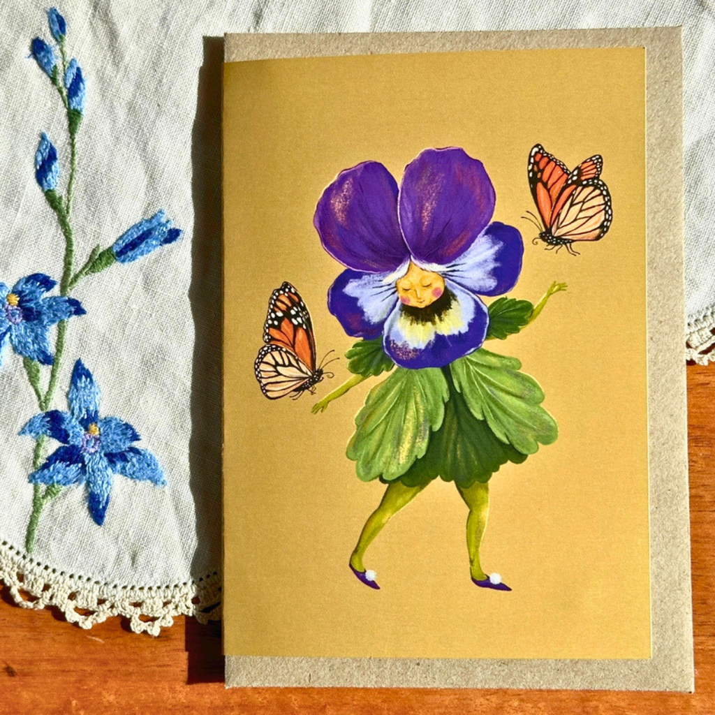 Pretty Pansy greeting card styled on a wooden surface with lace and blue flowers, featuring a cheerful pansy flower character accompanied by butterflies.