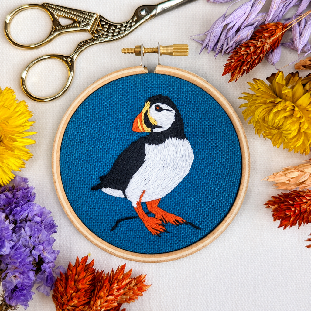 Finished puffin embroidery displayed with colourful threads, scissors and dried flowers.