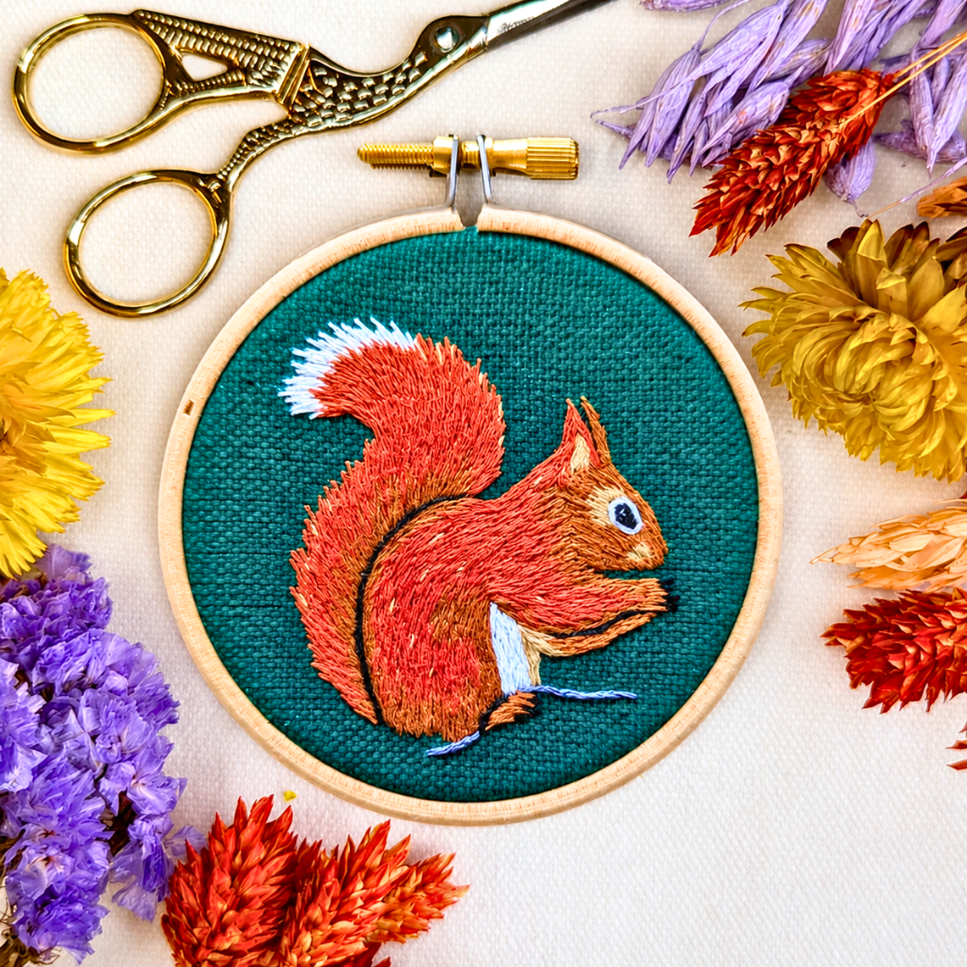 Finished red squirrel embroidery displayed with colourful threads, scissors and dried flowers.
