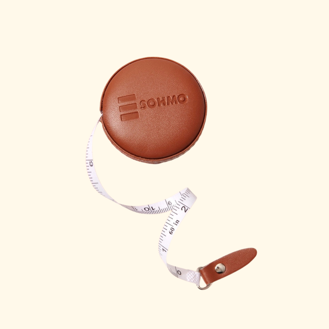 A brown leather retractable tape measure with a white measuring tape cord, shown on a light cream background. The brand name 'SOHMO' is embossed on the top of the circular case.
