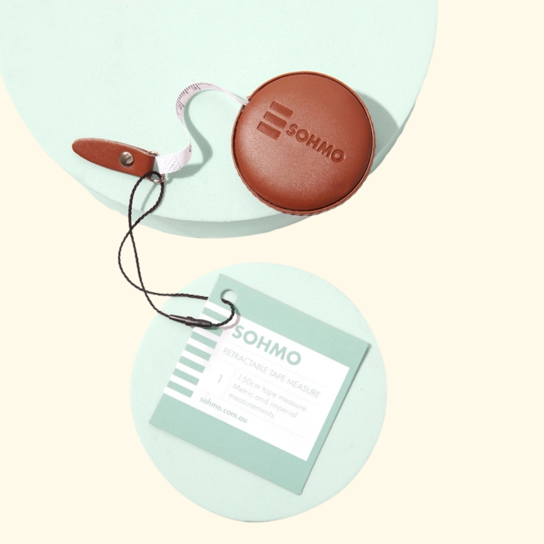 A round brown leather-like SOHMO tape measure with a retractable white tape arm, shown above a matching branded hangtag on a pale cream background with mint circular props.