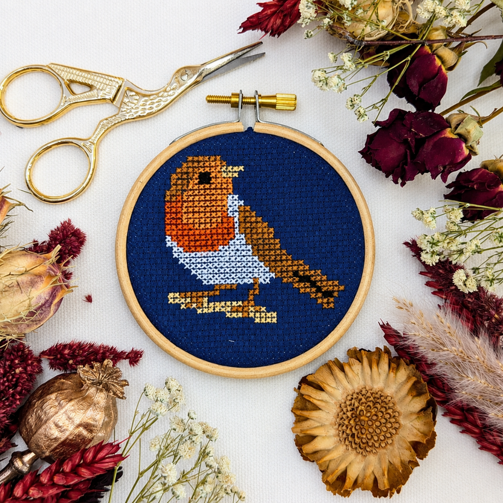 Finished robin cross stitch displayed in a wooden hoop with autumn leaves, seed pods and gold embroidery scissors.