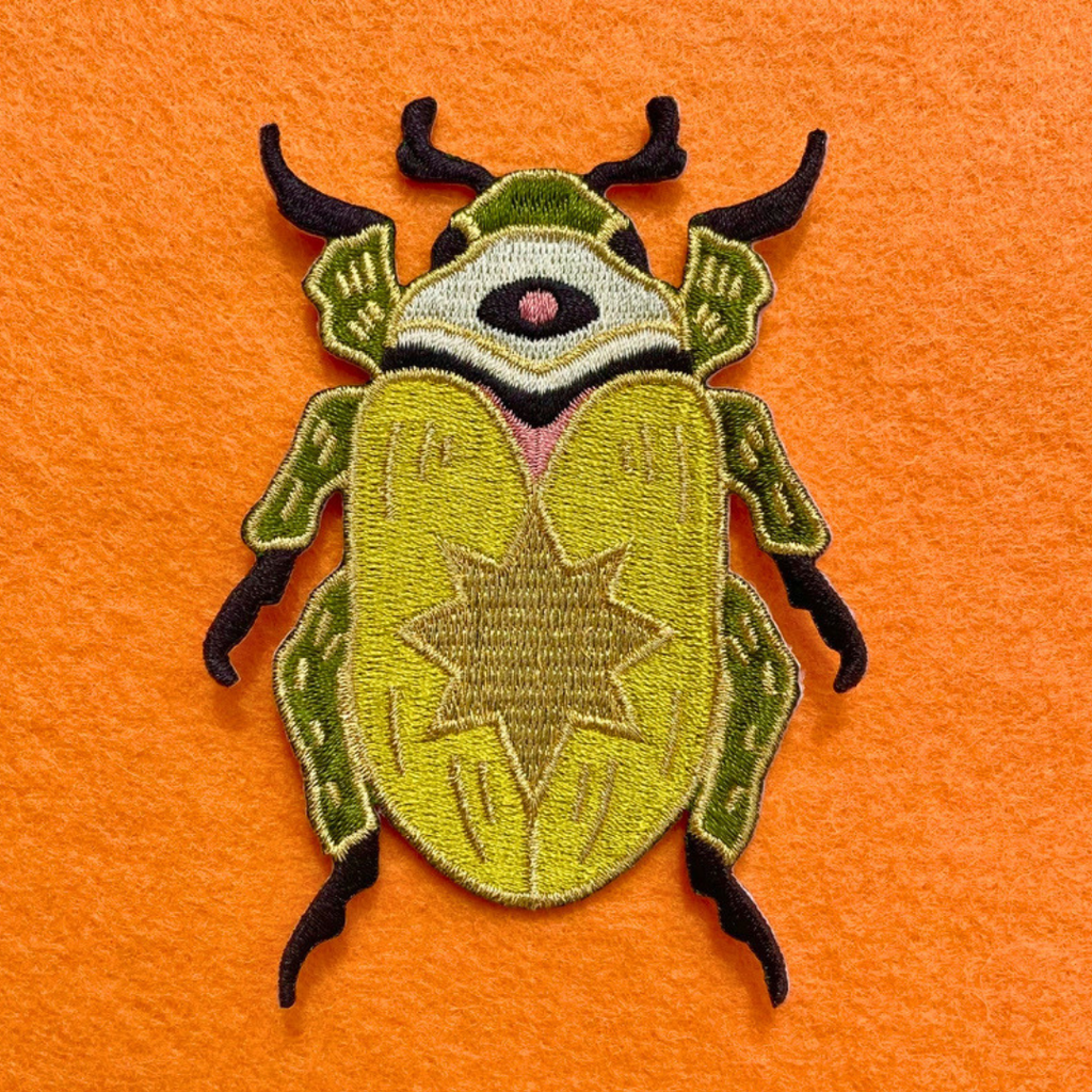 Close-up of Ashlea Bechaz Illustration scarab beetle embroidered iron on patch featuring green and yellow stitching against a bright orange background.