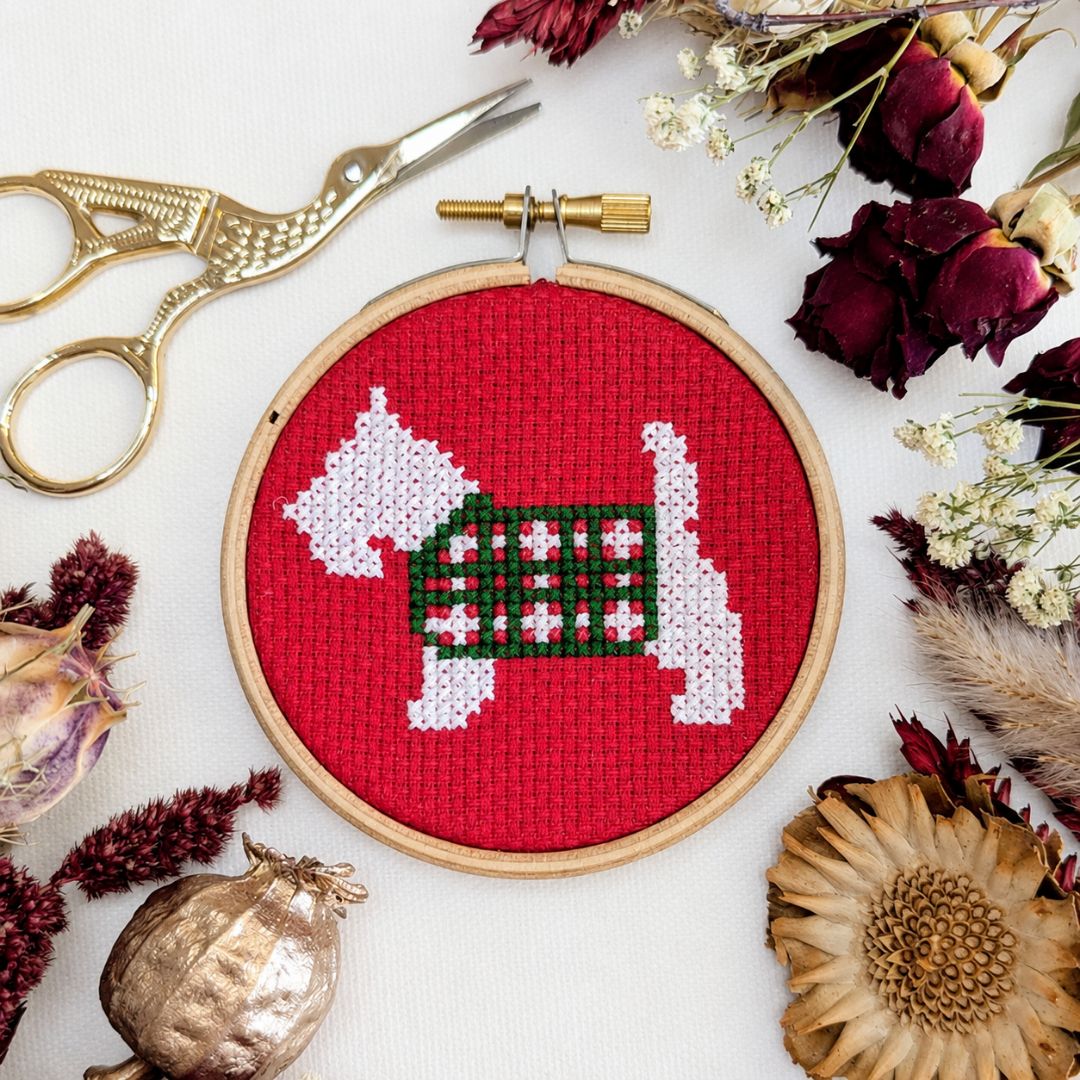 Finished Scottie dog cross stitch displayed in a wooden hoop with flowers, leaves and decorative craft accessories.